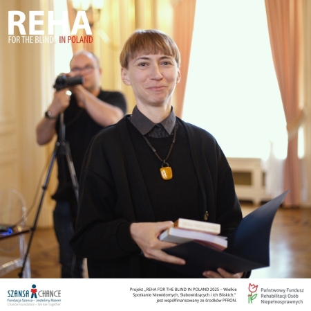 Na zdjęciu osoba uśmiechnięta, stojąca w eleganckim wnętrzu z dużym oknem i zasłonami w tle. Trzyma w rękach otwartą teczkę oraz kilka książek. Ubrana jest w ciemną odzież i naszyjnik z dużym bursztynowym wisiorkiem. W lewym górnym rogu widnieje napis „REHA FOR THE BLIND IN POLAND”, a na dole zdjęcia znajdują się logotypy Fundacji Szansa – Jesteśmy Razem oraz Państwowego Funduszu Rehabilitacji Osób Niepełnosprawnych.