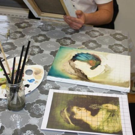 Widok na stół - reprodukcja obrazu oraz jego kopia we wstępnej fazie; malarska paleta, pędzle.