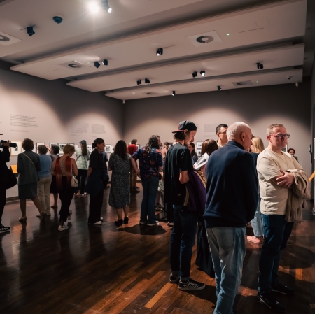 A wide shot of the interior of a crowded art gallery with graphite-colored walls and dark wooden floors. Many people are looking at framed photographs hanging along the walls, illuminated by ceiling spotlights. Some of the exhibits have descriptions placed next to them.
