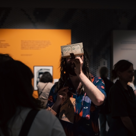 A man with dreadlocks, wearing a colorful shirt with an eye pattern, holds a vintage stereo viewer with a stereo card inserted into it in front of his eyes, looking toward an orange display panel in the background. Other visitors are visible as blurred figures around him.