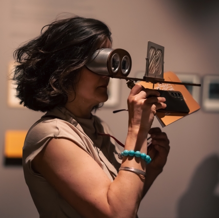 A middle-aged woman with dark, curly hair and a turquoise beaded bracelet is looking through an unusual stereo viewer with metal tubes for stereo cards, illuminated by a bright light from above. In her other hand, she is holding another pair of glasses and a piece of paper.