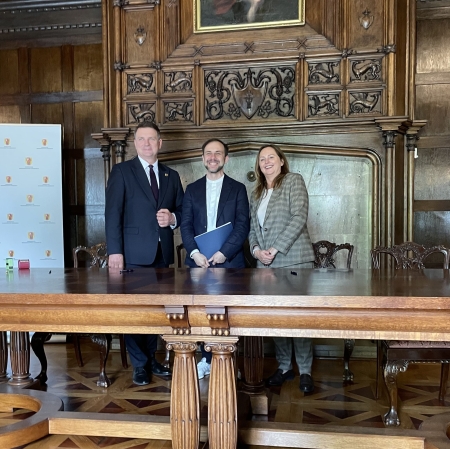 Dwóch mężczyzn i kobieta pozują na tle kominka oraz wiszącego nad nim obrazu. Przed pozującymi duży drewniany stół.