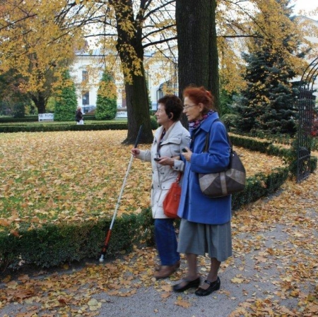 Dwie kobiety idą bardzo blisko siebie alejką wśród drzew. Jedna z kobiet ma słuchawkę w uchu, a w dłoni trzyma laskę.