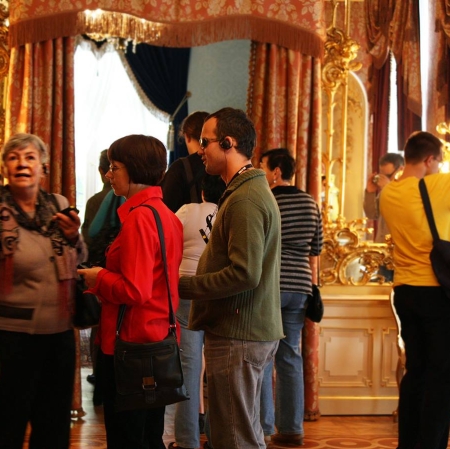 Kilka osób dorosłych we wnętrzu z dużymi lustrami. Mężczyzna na pierwszym planie ma w uchu słuchawkę i ciemne okulary na oczach.