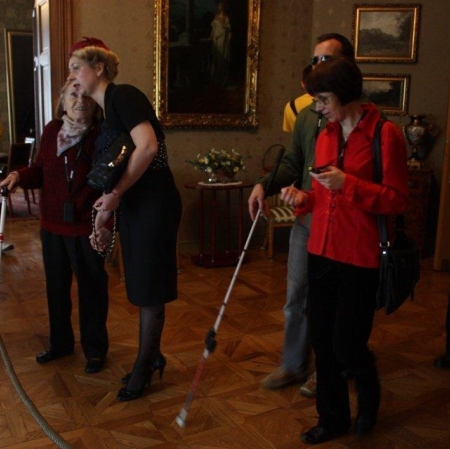 Mężczyzna w ciemnych okularach dotyka w rękawiczkach świecącego kinkietu przy lustrze.