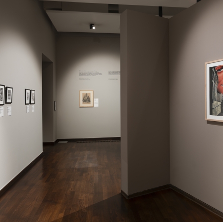 A section of the museum exhibition featuring a hallway leading to the next room. Framed photographs and prints hang on the walls, and in the foreground on the right is a large, colorful photograph depicting a section of a city billboard featuring a woman’s face. The entire space features a minimalist design with spot lighting.