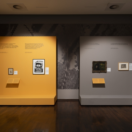 The interior of the exhibition hall features two displays on raised exhibition walls—one orange and the other gray. The walls showcase photographs, prints, and paintings accompanied by descriptions and quotes. The dark wooden floor and subdued lighting emphasize the space’s museum-like character.