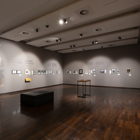 A wide view of the exhibition hall for “The School of Seeing: The City.” Photographs, prints, and curatorial texts are displayed on the walls, and in the center there is a display case and a bench for visitors. The space is lit by ceiling spotlights and features a dark wooden floor and light gray walls.