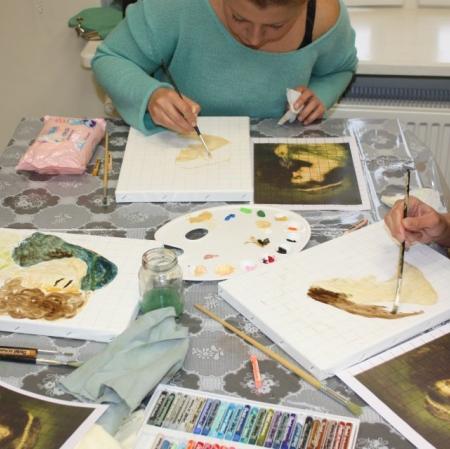 Zbliżenie na dłonie uczestników oraz powstające prace malarskie. Na stole paleta malarska, pędzle, pastele.