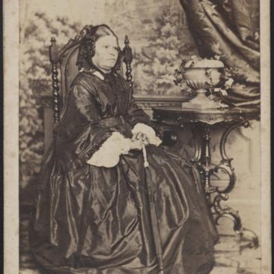 Czarno-biała fotografia kobiety w bogato zdobionym stroju, siedzi na krześle przy drewnianym stole. 