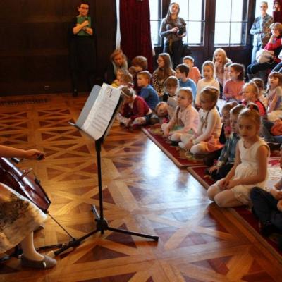 Zdjęcie: dziewczynka po lewej siedzi na krześle i trzyma wiolonczelę. Przed dziewczynką stojak na partyturę. Za partyturą na podłodze siedzi wiele dzieci. 