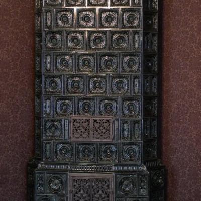 Ciemnobrązowy pie kaflowy, jego kafle są bogato ozdobione. Stoi w rogu na tle wzorzystej purpurowej tapety.