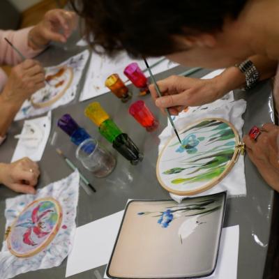 Trzy kobiety pochylające się nad obrazkami, które malują przy pomocy farb akwarelowych, na pracach widoczne kwiaty