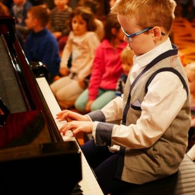 Zdjęcie: chłopczyk gra na fortepianie. W tle na podłodze siedzą dzieci.