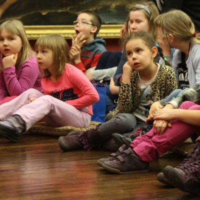 Zdjęcie przedstawia liczną grupę dzieci siedzących na drewnianej podłodze kierujących wzrok w jednym kierunku.