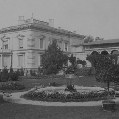 Czarno-biała fotografia ogrodu, na pierwszym planie fontanna otoczona kwiatami.