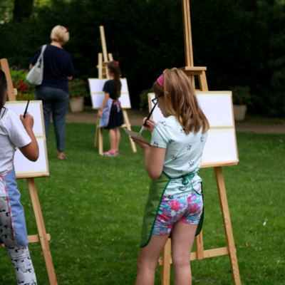 Dwie dziewczynki odwrócone tyłem, malują na kartonach umieszczonych na sztalugach
