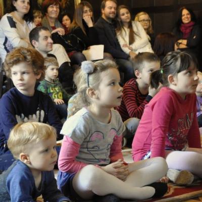 Zdjęcie: Liczna grupa dzieci siedzi na podłodze w dużej sali. Wszyscy patrzą w jednym kirunku.