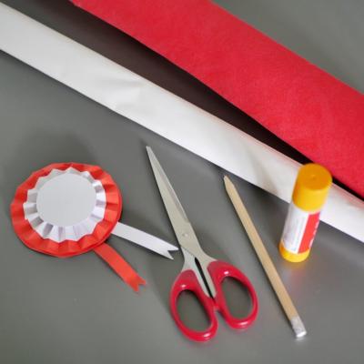 Na szarym tle nożyczki, ołówek, klej w sztyfcie oraz biało czerwony kotylion, obok biały, czarny i czerwony papier.