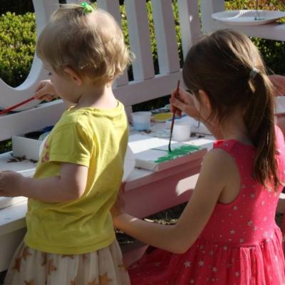 Zdjęcie: dwie dziewczynki przed ławeczką, na której leżą kartki.  Dziewczynki trzymają pędzle w dłoniach.