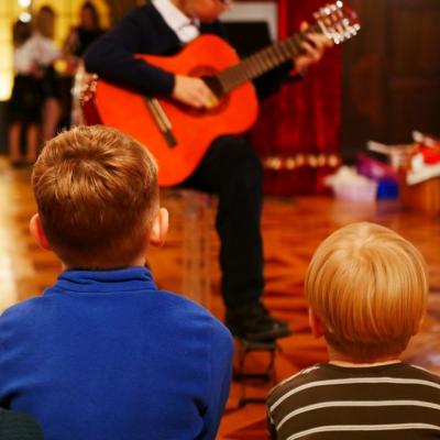 Zdjęcie: chłopczyk siedzi na krześle z gitarą we wnętrzu. Przed chłopcem na podłodze siedzi dwoje dzieci. 