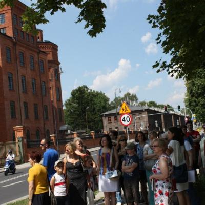 Zdjęcie: liczna grupa dorosłych stoi na chodniku. W tle fragment dawnej Nowej Przędzalni Scheiblera
