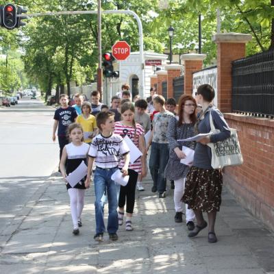 Zdjęcie przedstawia liczną grupę dzieci idącą wzdłuż ulicy. Z przodu grupy młoda kobieta.
