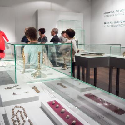 Zdjęcie przedstawia wnętrze ekspozycji wystawowej, w której stoi kilka osób.