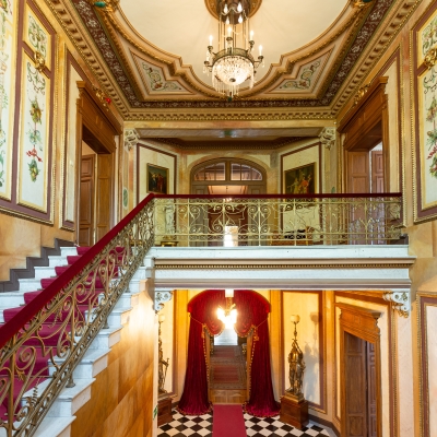 Elegancki, pałacowy westybul z marmurowymi schodami wyłożonymi czerwonym dywanem, ozdobną balustradą i kryształowym żyrandolem.