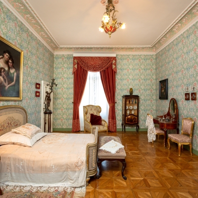 An elegant, historic bedroom with floral wallpaper, an antique bed, red window drapes, vintage furnishings, and a parquet floor.