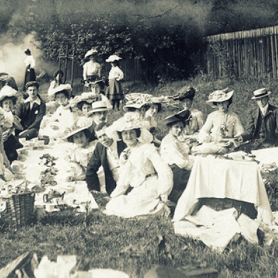 Czarno-białe zdjęcie przedstawia liczną grupę kobiet siedzących na trawie ubrane w długie białe suknie. Kobiety oraz nienieliczni mezczyźni mają kapelusze na głowach.