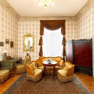 A classic, elegant boudoir featuring floral wallpaper, mustard-yellow velvet armchairs and sofa, brown window drapes, a ornate gold-framed mirror, and an oriental floor rug.