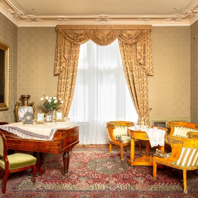 An elegant 19th-century family salon featuring patterned gold wallpaper, a grand piano, striped antique armchairs, framed portraits, and an ornate rug.