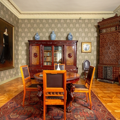 A historic home library with a large wooden bookcase, a ornate tiled stove, a classical bust on a pedestal, and a central table with leather chairs.