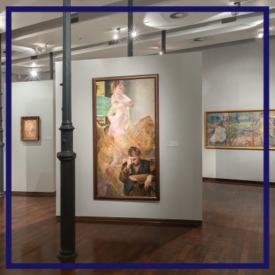 Zdjęcie oprawione w granową ramę przedstawia wnętrze galerii. Na białych śniach wiszą obrazy.