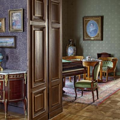 Zdjęcie przedstawia wnętrza. Ujęcie fragmentu pokoju, w którym po lewej stoi drewniana komoda, na ścianie nad komodą wiszą niewielkie obrazy w pozłacanych ramach. Otwatwarte drzwi do pokoju obok, w którym stoi fortepian, krzesło, fotel, kanapa i stolik