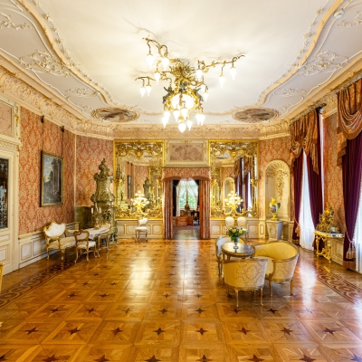 Bogato zdobiony, pałacowy salon lustrzany z parkietem we wzory, bogatymi sztukateriami na suficie, ozdobnym żyrandolem oraz dużymi lustrami w złoconych ramach. Przy oknach wiszą ciężkie, bordowo-złote zasłony, a wzdłuż ścian stoją stylizowane, złote meble.