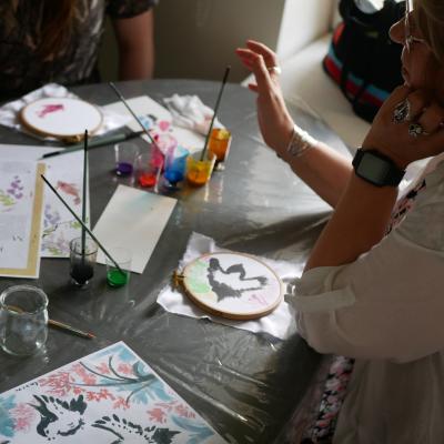 Zdjęcie przedstawia starszą obietę siedzącą przy stole, na którym leżą ilustrastracje, kubeczki z farbkami, pendzelki. Przed kobietą na stole leż niewielki drewniany tamborek z naciągniętą białą tkaniną. Na tkaninie namalowany biało-czarny ptak