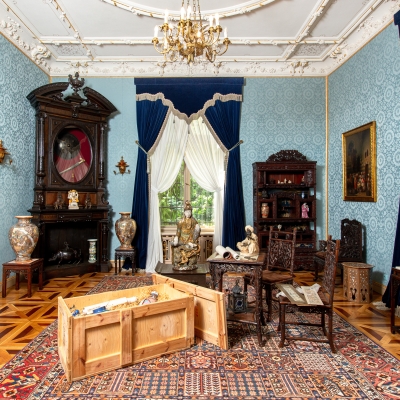 Elegancki salon w stylu orientalnym z niebieskimi tapetami, wzorzystym dywanem i ciemnymi, rzeźbionymi meblami. W pomieszczeniu znajdują się ozdobne wazy, figurki, kominek oraz otwarta drewniana skrzynia na pierwszym planie, a okna zdobią granatowe i białe zasłony.