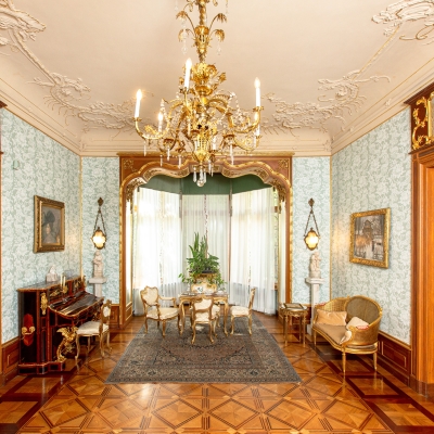 Jasny, elegancki salon z jaśminowo-niebieską wzorzystą tapetą, ozdobną sztukaterią na suficie oraz dużym złotym żyrandolami. W głębi znajduje się wnęka okienna ze stołem i krzesłami, po lewej stronie stoi zabytkowe pianino, po prawej mała kanapa, a po obu stronach pomieszczenia widoczne są masywne, drewniane drzwi.