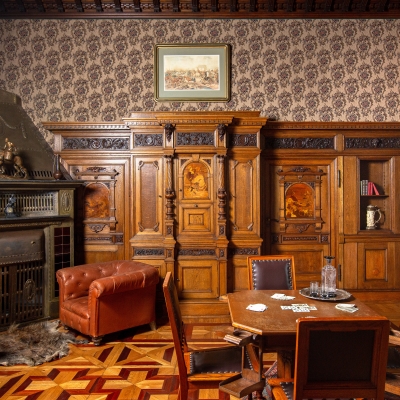 Klimatyczny salon myśliwski z wyrazistą, drewnianą boazerią i ozdobną tapetą. W centrum znajduje się ośmiokątny stół do gry w karty z krzesłami, po lewej stronie duży kominek i skórzane fotele, a w tle misternie rzeźbione meble oraz kasetonowy sufit.