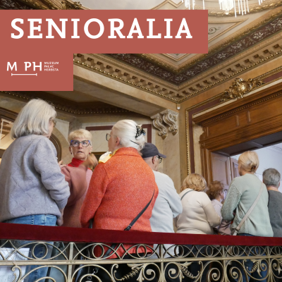 Seniorzy stoją obok siebie w przestrzebi zabytkowego pałacu, opierając się o poręcz.