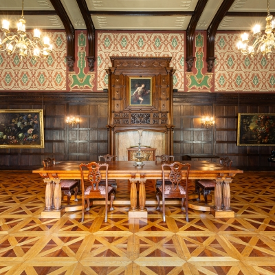 A grand ballroom featuring geometric parquet wood flooring, dark wood wall paneling, hanging chandeliers, framed paintings, and a long wooden conference table surrounded by carved chairs.