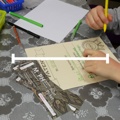 Zdjęcie przedstawia dłonie dziecka wypełniającego arkusz z napisem Patent, który leży na wzorzystym obrusie obok biletów do Muzeum Pałacu Herbsta. Na stole znajdują się również kolorowe mazaki oraz czysta kartka papieru.