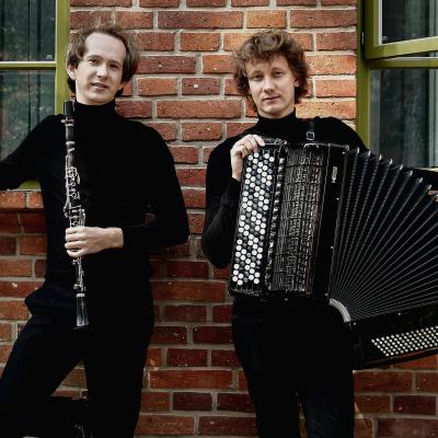 Dwóch mężczyzn. Jeden z klarnetem, drugi z akordeonem. Stoją na tle ściany z czerwonej cegły.