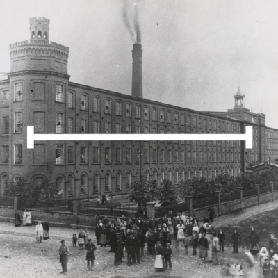 Czarno-białe zdjęcie przedstawia dużą ceglaną fabrykę z dwiema wieżami i wysokim kominem dymiącym na tle nieba. Przed budynkiem stoi tłum ludzi – mężczyzn, kobiet i dzieci – ubranych w stroje z przełomu XIX i XX wieku. Przez środek obrazu biegnie pozioma biała linia.