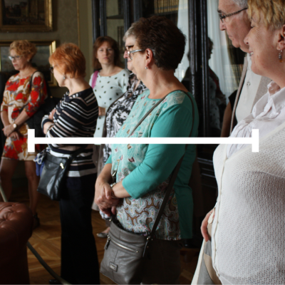 Grupa dorosłych osób zwiedzających wnętrze muzeum lub zabytkowego pomieszczenia. Wszyscy stoją w półokręgu, słuchając przewodnika. W centrum zdjęcia znajduje się pozioma, biała linia.