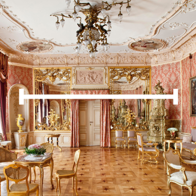 Bogato zdobione wnętrze pałacowe z ozdobnym żyrandolem, wzorzystą tapetą w kolorze łososiowym i kunsztownymi sztukateriami na suficie. Na środku zdjęcia znajduje się duża, biała, pozioma linia zakończona pionowymi kreskami. Pomieszczenie wyposażone jest w złocone meble, liczne lustra w ozdobnych ramach oraz dekoracyjny piec kaflowy po prawej stronie. Podłoga wykonana jest z jasnego parkietu ułożonego w geometryczne wzory
