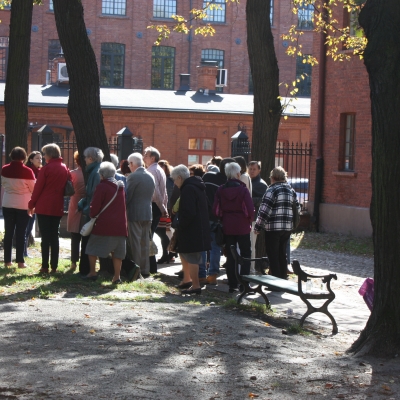Zdjęcie przedstawia grupę ludzi na spacerze. Ukazani tyłem na tle czerwonego, ceglanego budynku. Ich sylwetki widoczne są między drzewami.