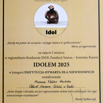 Dyplom w kolorze jasnego beżu z czarną ramką ozdobną, przyznany przez Fundację Szansa – Jesteśmy Razem w ramach regionalnego Konkursu IDOL.  Tekst główny informuje, że:  Za zajęcie I miejsca w regionalnym Konkursie IDOL Fundacji Szansa – Jesteśmy Razem IDOLEM 2025 w kategorii INSTYTUCJA OTWARTA DLA NIEWIDOMYCH został/została Muzeum Pałac Herbsta – Oddział Muzeum Sztuki w Łodzi.